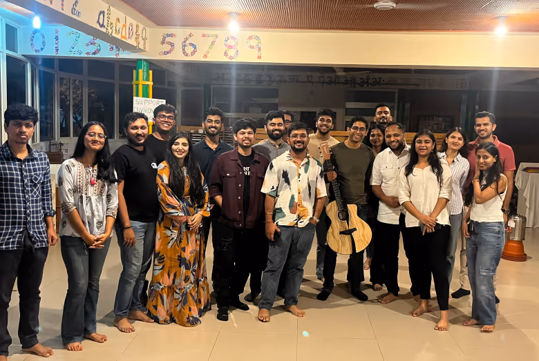Group of young adults standing barefoot indoors, one holding an acoustic guitar, smiling at the camera.