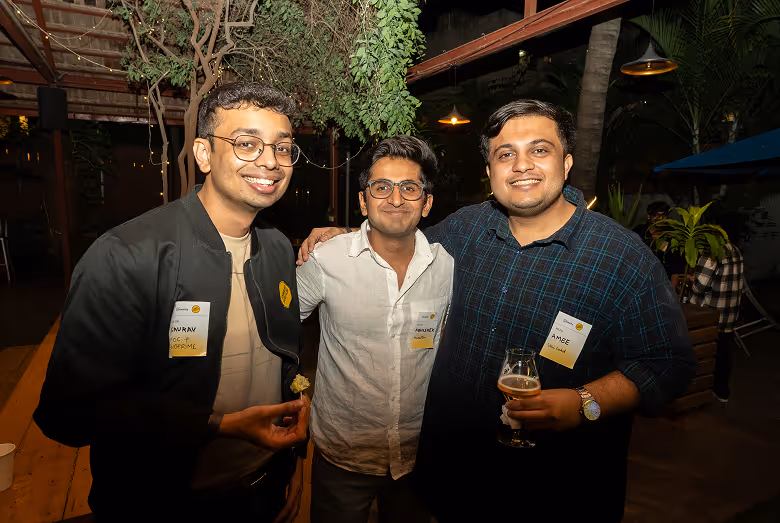 Three men smiling indoors at a social event, two wearing glasses and name tags, one holding a drink.