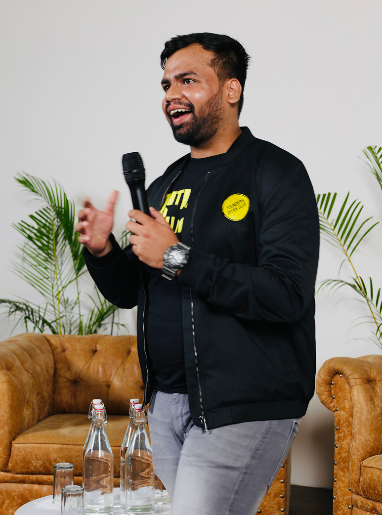 Man in black jacket speaking into a microphone beside brown leather sofas and plants.