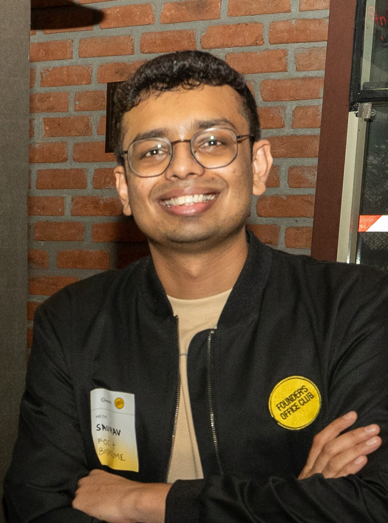 Smiling man wearing glasses and a black jacket with patches stands against a brick wall.