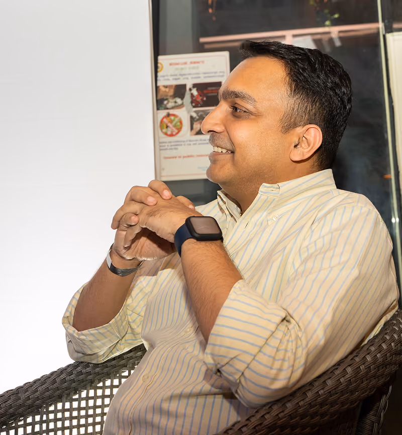 Man in striped shirt sitting on wicker chair with hands clasped, smiling and looking right.