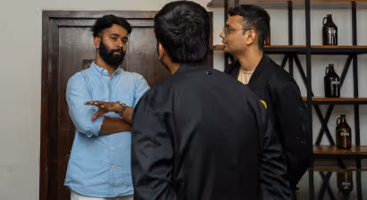 Three men in conversation; one wearing blue shirt with arms crossed, others facing him in black jackets.