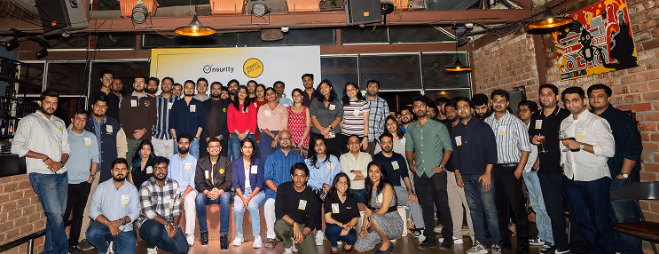 Group photo of diverse people posing indoors in front of a screen with logos on a brick wall background.