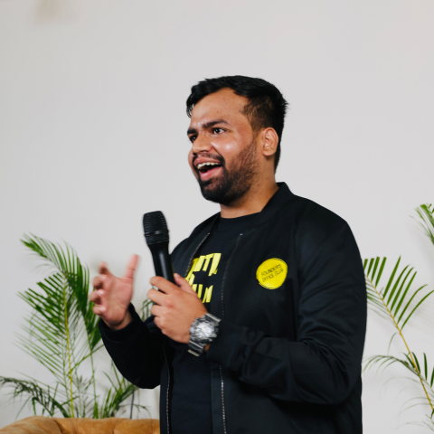 Man speaking into a microphone with a smile, standing in front of a couch and green plants.