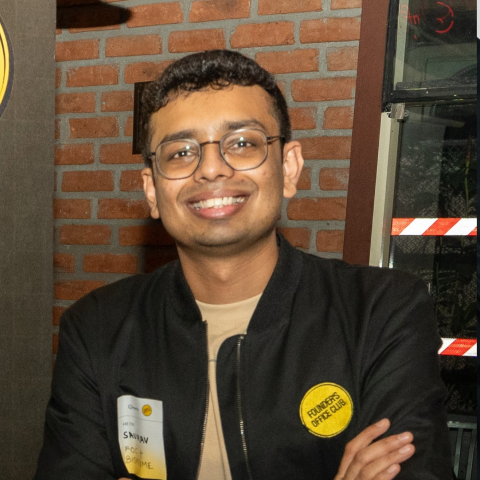 Smiling man wearing glasses and a black jacket standing with arms crossed against a brick wall.
